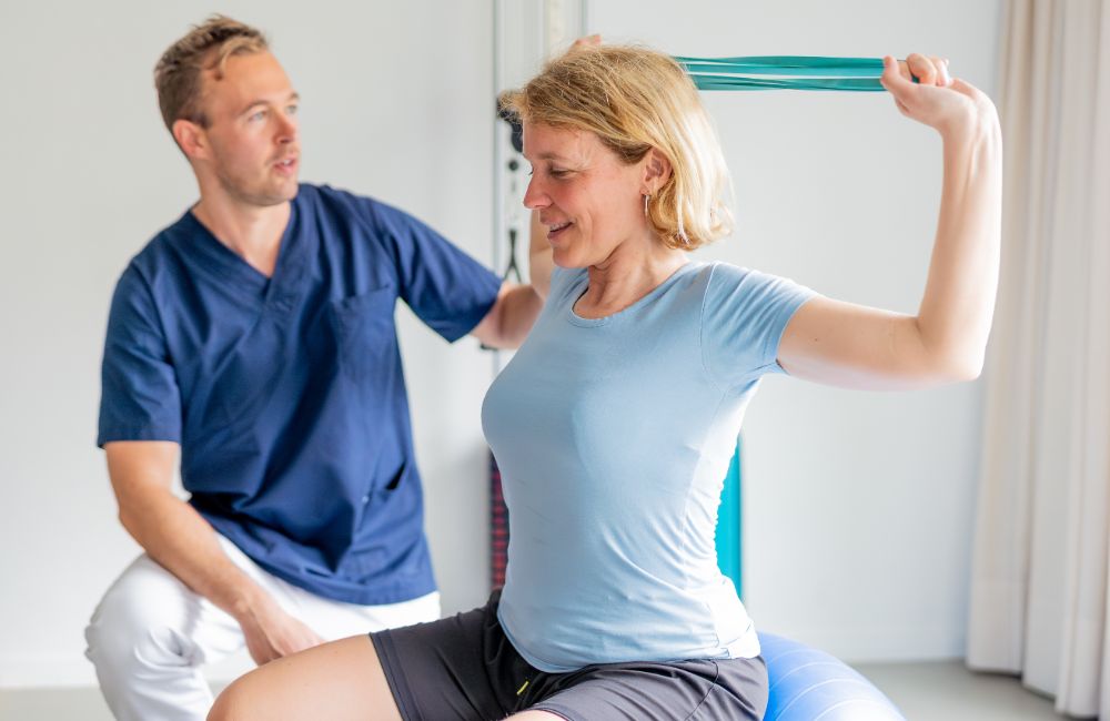 Physical therapist assisting patient exercising with resistance band on fitness ball. Patient doing exercises with resistance band assisted by physical therapist for rehabilitation